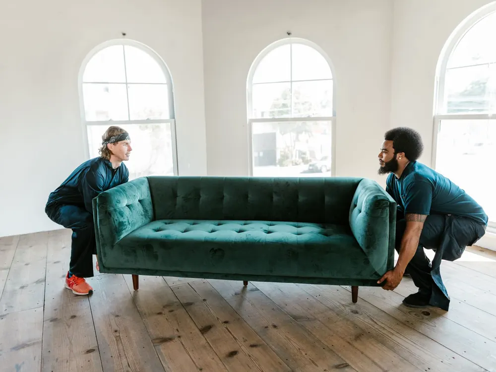 Two movers carrying a green sofa across an empty apartment