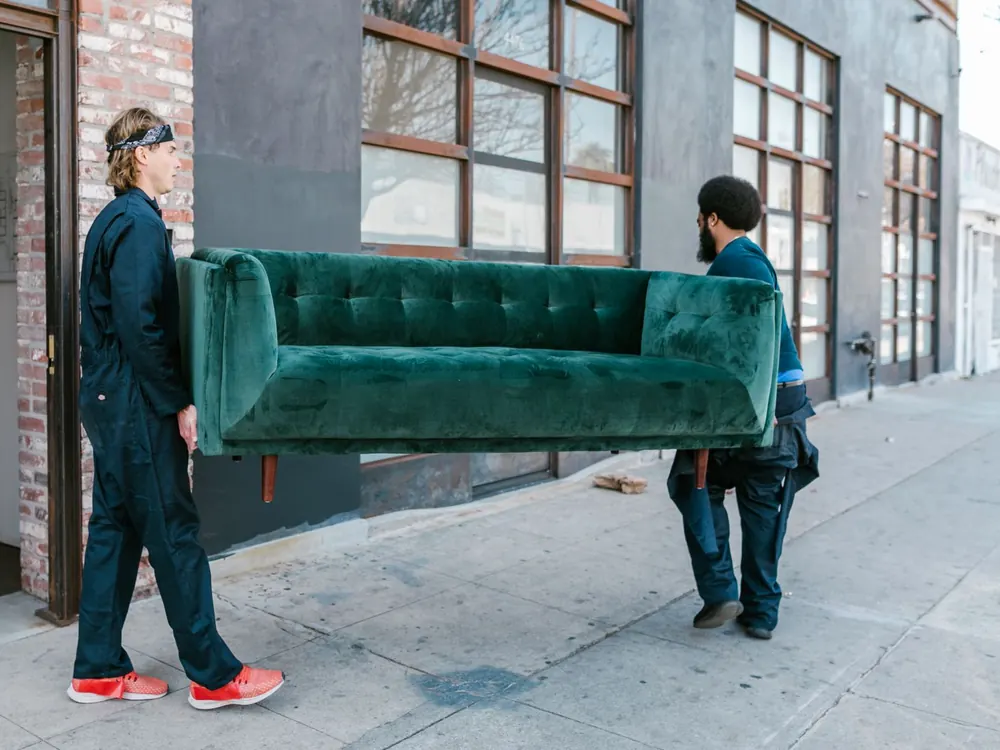 Two movers carrying a green sofa along the street to the truck